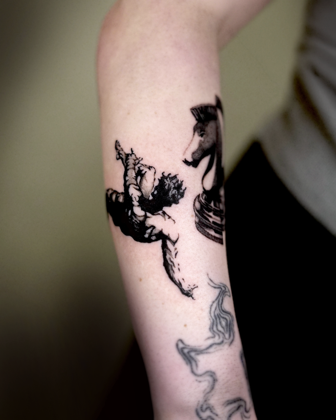 Black and grey woodcut tattoo on forearm: knight on a rearing horse with a small smoke-line figure below