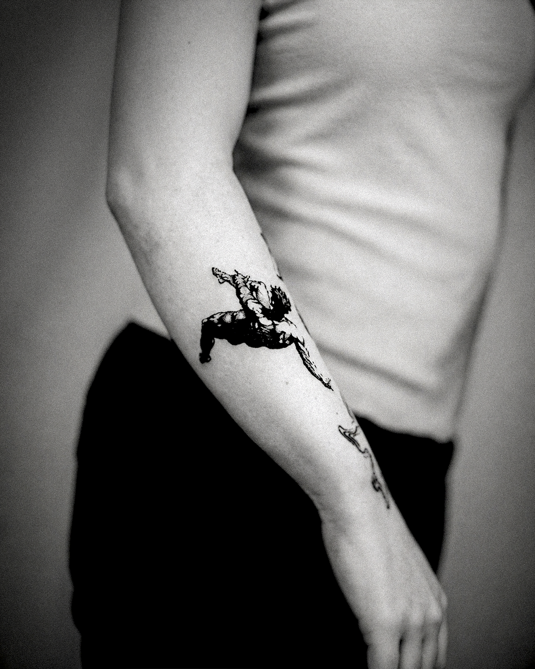 Black and grey woodcut tattoo on inner forearm: a falling winged figure mid-plunge, dynamic linework against bare skin
