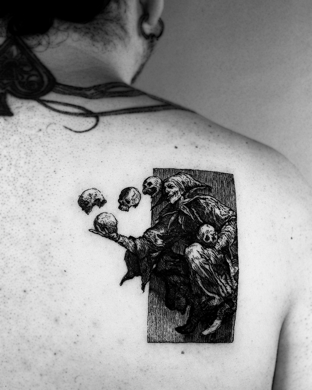 Black and grey woodcut back piece: a hooded jester holding a skull, three more skulls floating around him, framed inside a tall rectangle